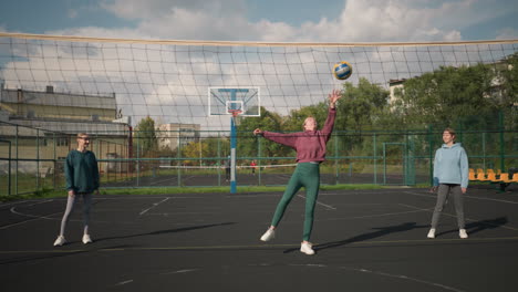 volleyball players standing in court as lady in green leggings prepares to slam volleyball over net with basketball hoop in background, captured in outdoor sports arena with greenery and buildings