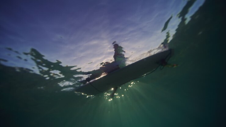 Backlit silhouette of surfer sitting on longboard, view from underwater