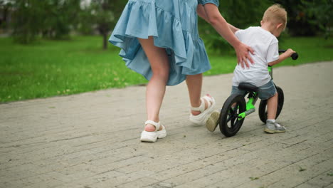 a caring mother guides her little boy by pushing him gently as he starts riding his bicycle along an interlocked pathway, he begins to ride faster, another child rides in the background