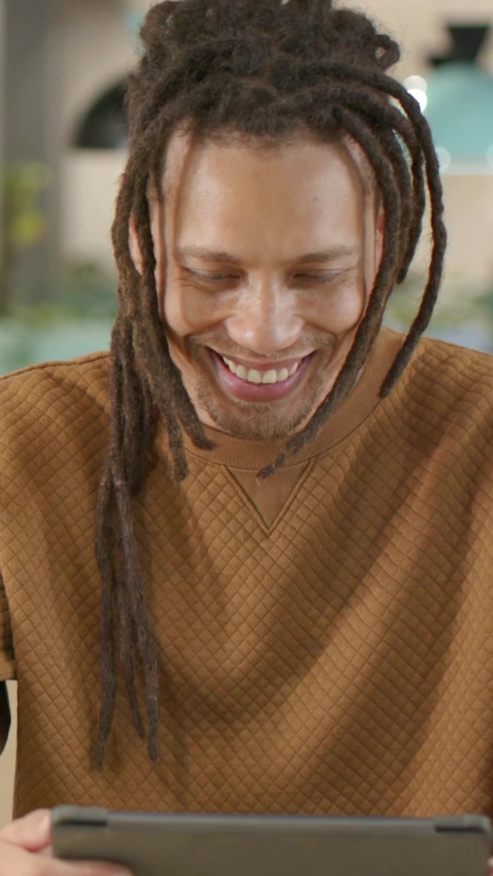 Video verticale: Uomo sorridente con maglione marrone che reagisce al tablet in un caffè, toccando lo schermo, con braccialetti e un anello.