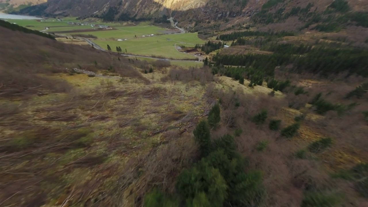 Højhastigheds FPV-drone flyver ned fra en stejl bjergskråning ind i en grøn alpedal med sneklatter og snoede veje