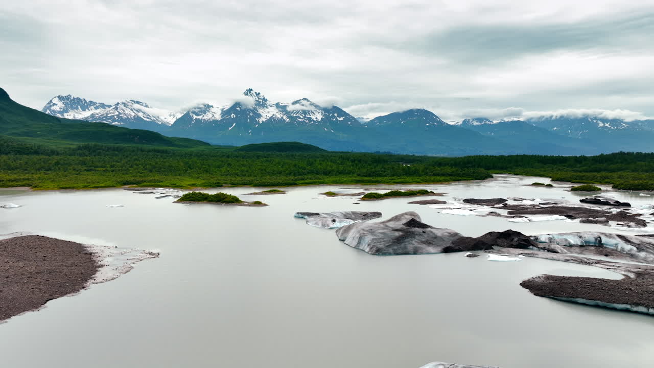 Drone footage over the river with melting ice pieces. Approaching green waterfront surrounded by stunning snow-capped mountains. Alaska, USA