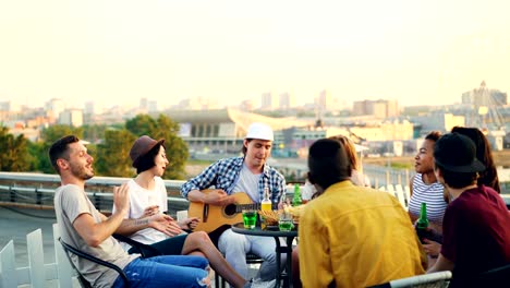 students are having fun on rooftop playing the guitar, singing and dancing sitting at table on rooftop enjoying free time. modern lifestyle and musical instruments concept.