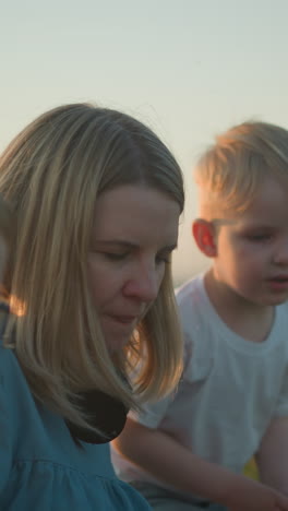 una madre con una bata azul sentada en un campo cubierto de hierba, sosteniendo una computadora portátil. sus dos hijos pequeños, uno de amarillo y el otro de blanco, se paran y se sientan a su lado, observando con curiosidad su trabajo