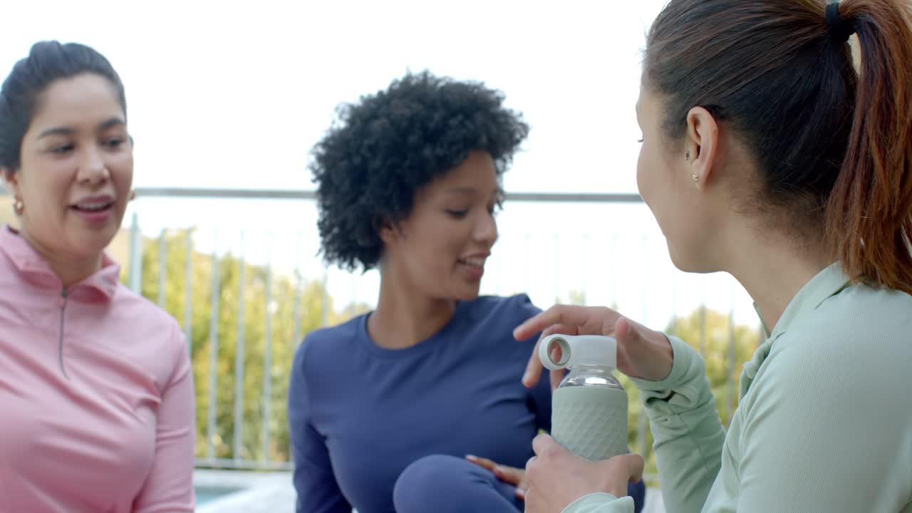 After yoga, Diverse female friends in activewear reaching for water bottle and hydrating on terrace