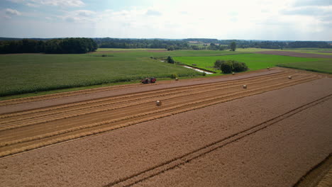 aerial backwards shot of tractor harvesting corn of farm field in countryside at summer season - forest woodland in backdrop