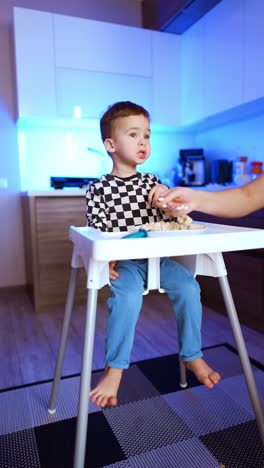 lovely baby boy sitting at high chairs having meals. mom's hand picks some porridge from a plate with a spoon. vertical video.