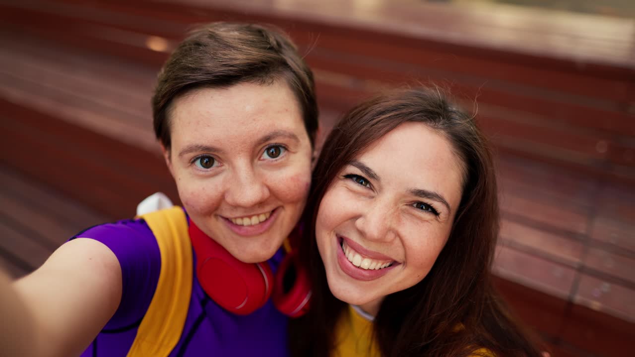 retrato de una chica morena con un suéter amarillo y una chica con un corte de pelo corto en un top púrpura y auriculares rojos que se toman una selfie en un banco marrón en un skatepark en verano