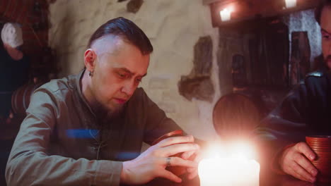 man with fellow drinks in medieval tavern. sad guy with goatee looks inside empty clay mug sitting by friend in ancient inn. guest has no money for beer