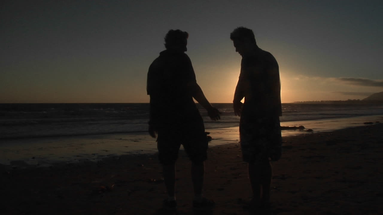 바람 부는 해변에서 두 남자의 실루엣이 손을 잡고 있다