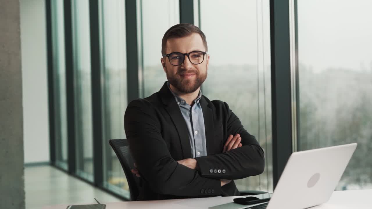 uomo sorridente in abiti da lavoro e occhiali seduto su una sedia davanti a un computer portatile guardando la telecamera e sorridente
