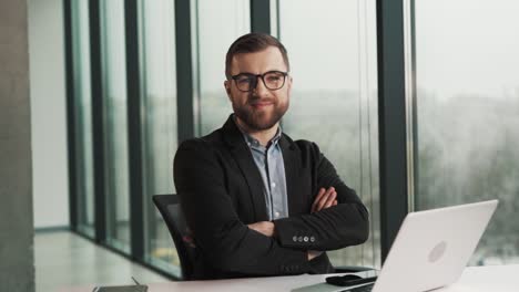 lächelnder mann in geschäftskleidung und brille, der auf einem stuhl an einem laptop sitzt, in die kamera schaut und lächelt