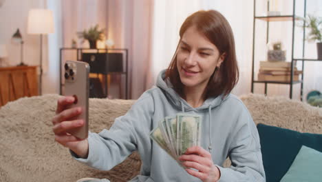 smiling woman blogger influencer recording story on smartphone sitting on sofa showing money at home