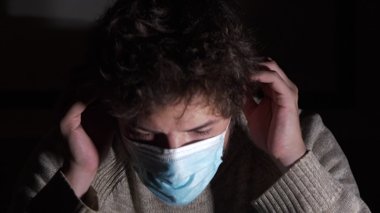 暗闇でカメラを見ながらコロナウイルス用サージカルマスクを装着する若い白人男性