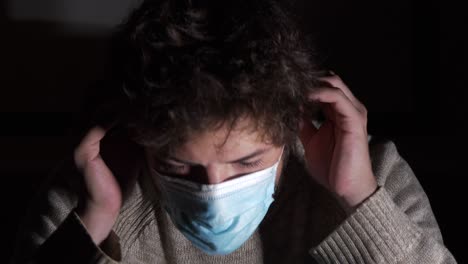 Young-Caucasian-male-putting-on-corona-virus-surgical-mask-looking-at-camera-in-darkness