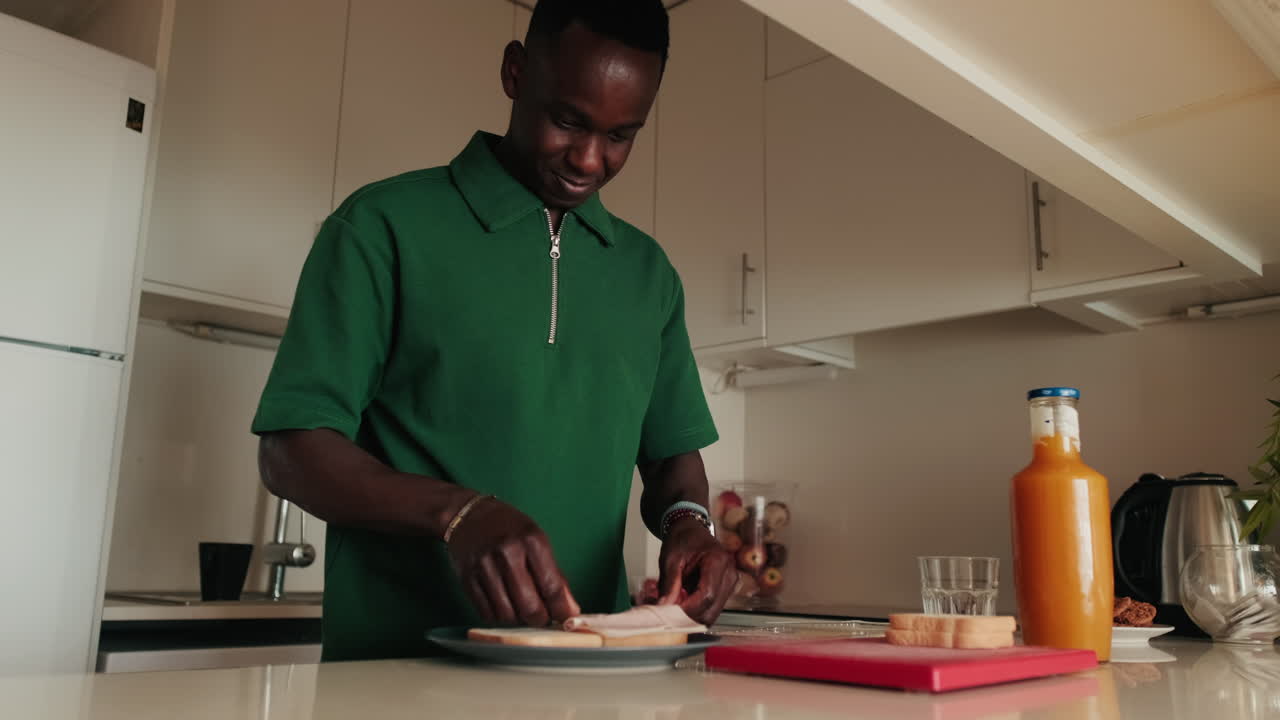 Smiling Young Man Prepares a Ham Sandwich in His Kitchen