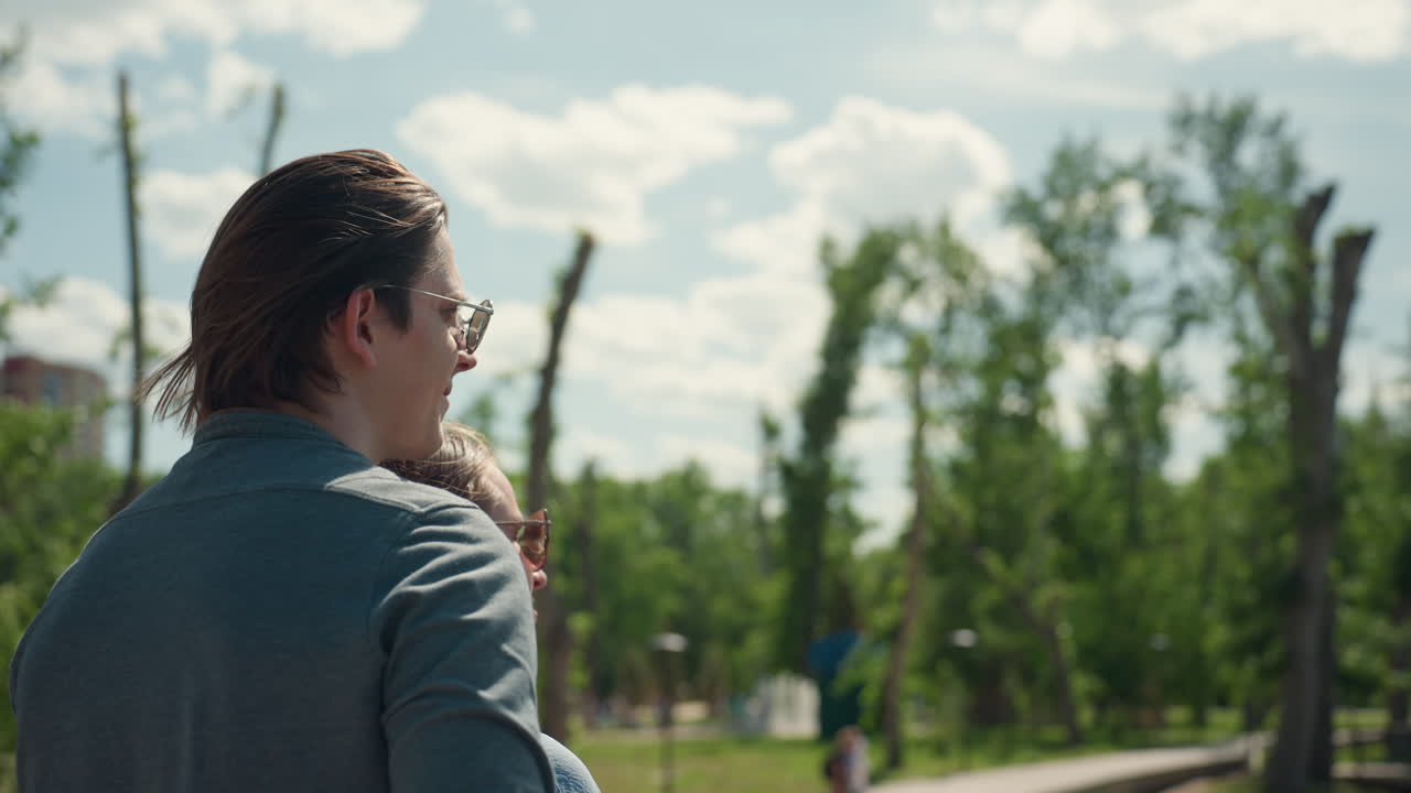 Un hombre blanco señala el horizonte de un lago desde un banco con el brazo extendido. Las gafas de sol reflejan el cielo, su pelo ondea al viento y su gesto guía la mirada hacia el agua y los barcos a lo lejos, en un ambiente contemplativo en un parque exuberante.