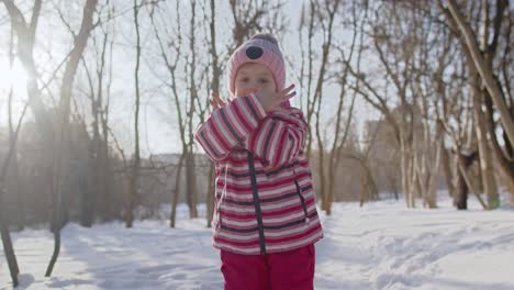 Joyful-little-child-girl-looking-at-camera,-smiling,-dancing-on-snowy-road-in-winter-park-outdoors