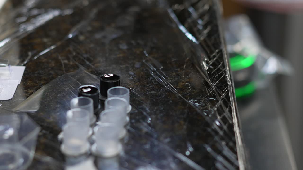 Tattoo artist gloved hand dipping machine or gun needle tip in black ink cups on sanitized cling wrap work shop station, with green glowing power supply in background