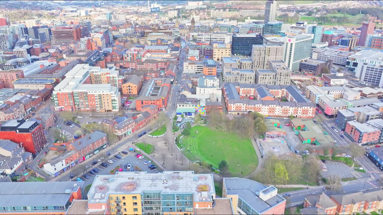 Drone shot captures Sheffield city center showing Devonshire Green park, skatepark, surrounding modern apartments, older brick buildings, and city skyline under soft daylight, aerial view