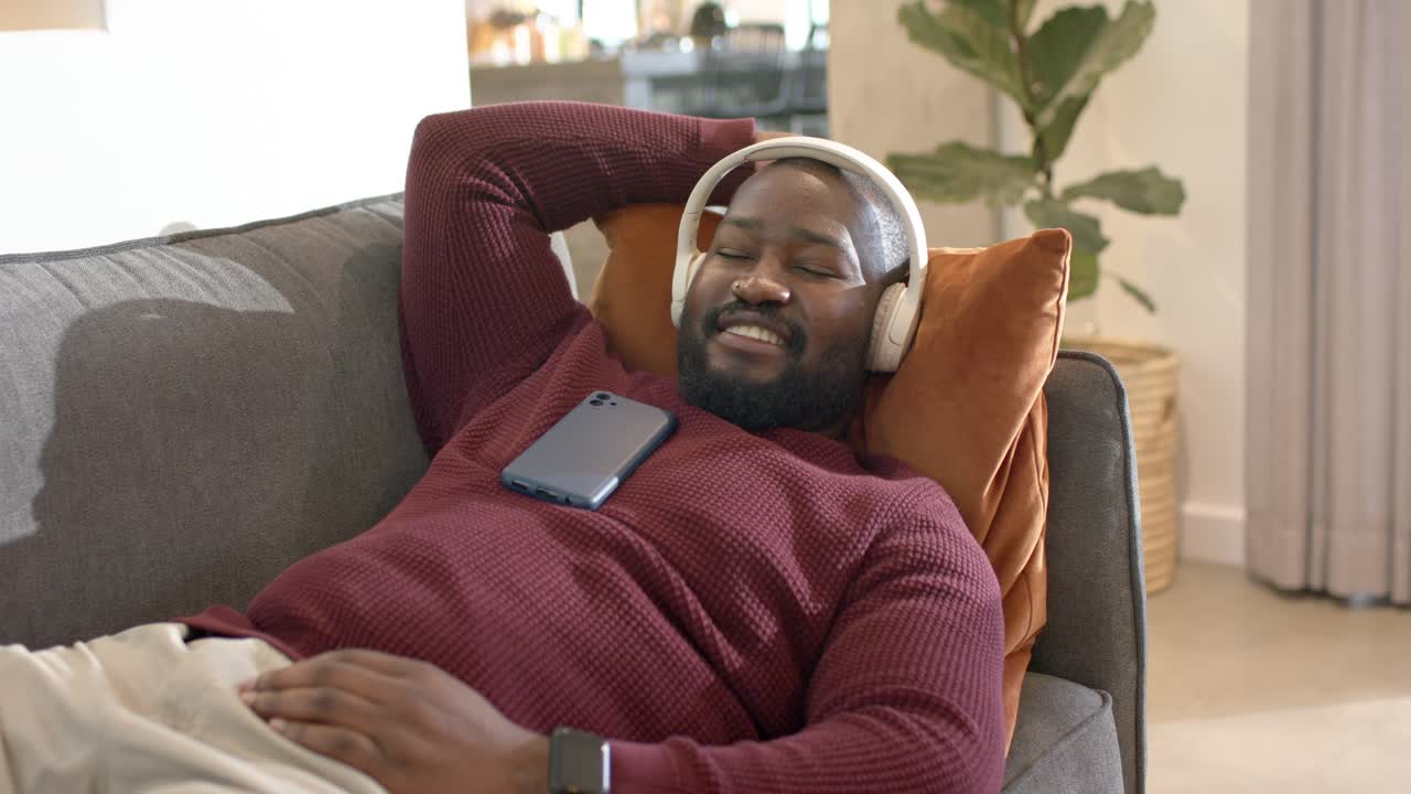 African American man lifting phone and scrolling on sofa then leaning back and listening to music