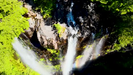 aerial dolly out of three waterfalls falling into rocky natural pool forming a rainbow surrounded by dense green forest