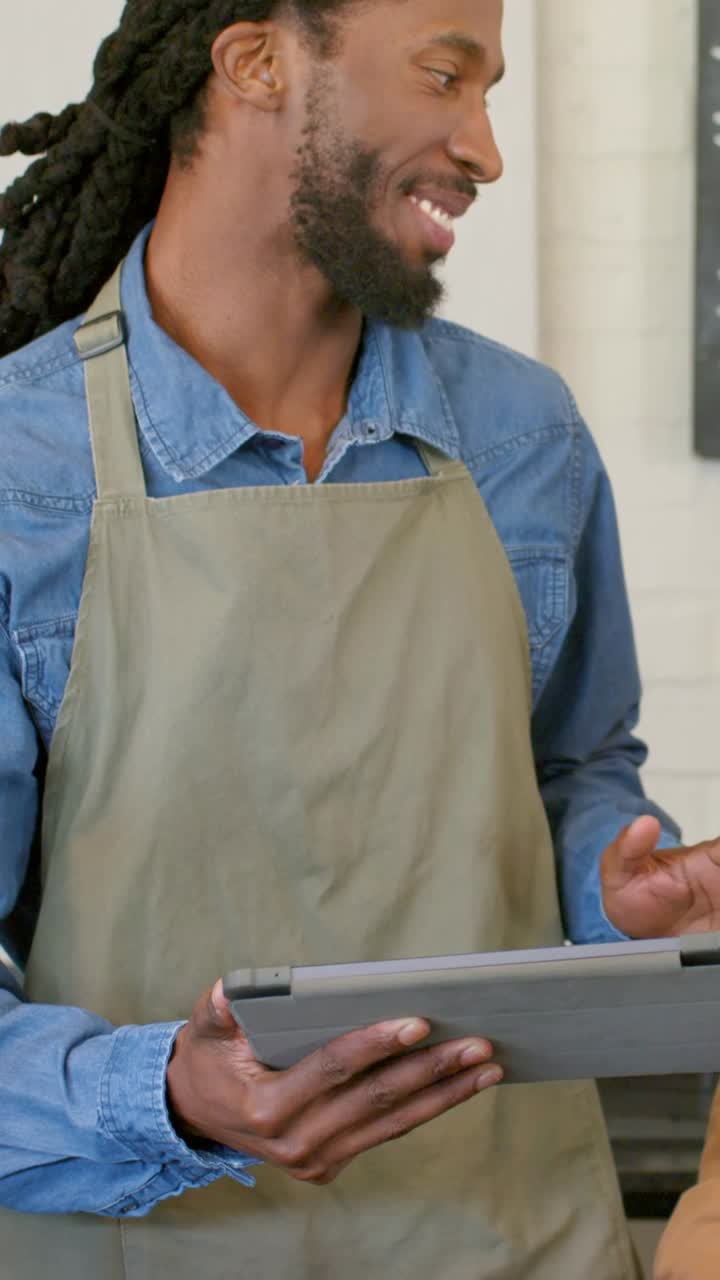 Vertical video: Responding to customer, barista wearing khaki apron holding tablet at cafe counter