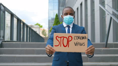 portrait of caucasian elegant young african american man in facial mask showing stop coronavirus" signboard in the street"