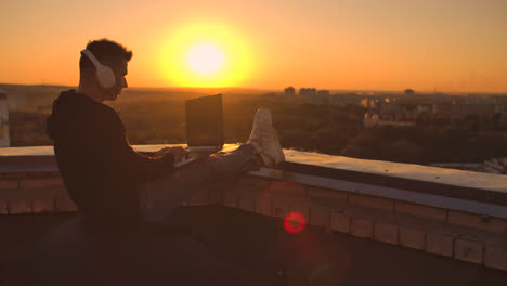 Ein-Mann-Mit-Kapuzenpullover-Sitzt-Auf-Dem-Dach-Und-Hört-Musik-Mit-Kopfhörern-Und-Tippt-Auf-Der-Tastatur-Eines-Laptops.-Laufen-Bei-Sonnenuntergang.-Freiberufler-Arbeitet-Bei-Sonnenuntergang-Mit-Einem-Laptop