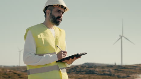 advancing clean and renewable energy, an engineer uses software technology on a tablet to audit wind turbines in a field of green energy generators on a sunny day while wearing a white helmet