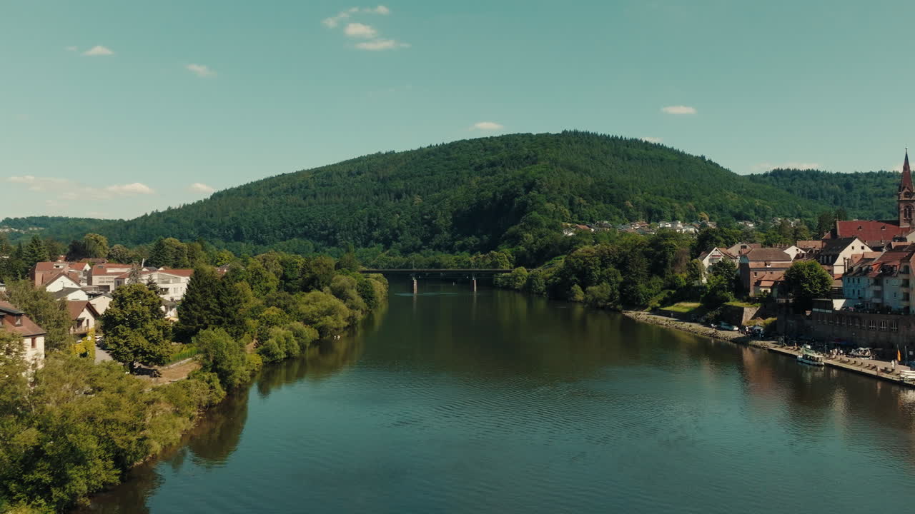 Luftfoto i lav højde af en ren og rolig flodkanal med smukke grønne bjerge og en bro i baggrunden samt bebyggelser på begge flodbredder