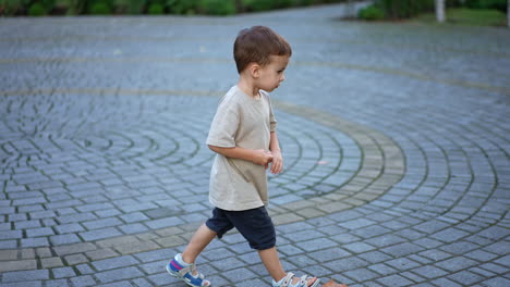 little kid in t-shirt and shorts stands in the middle of paved square. lovely baby boy comes up to his scooter and rides it slowly.