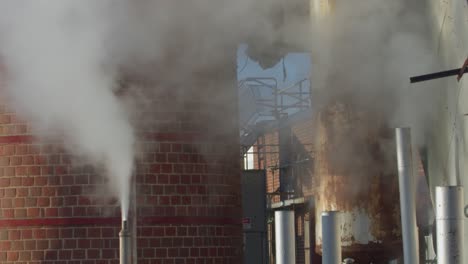 Billowing-smoke-from-industrial-pipes-against-a-building-with-clear-skies,-urban-setting