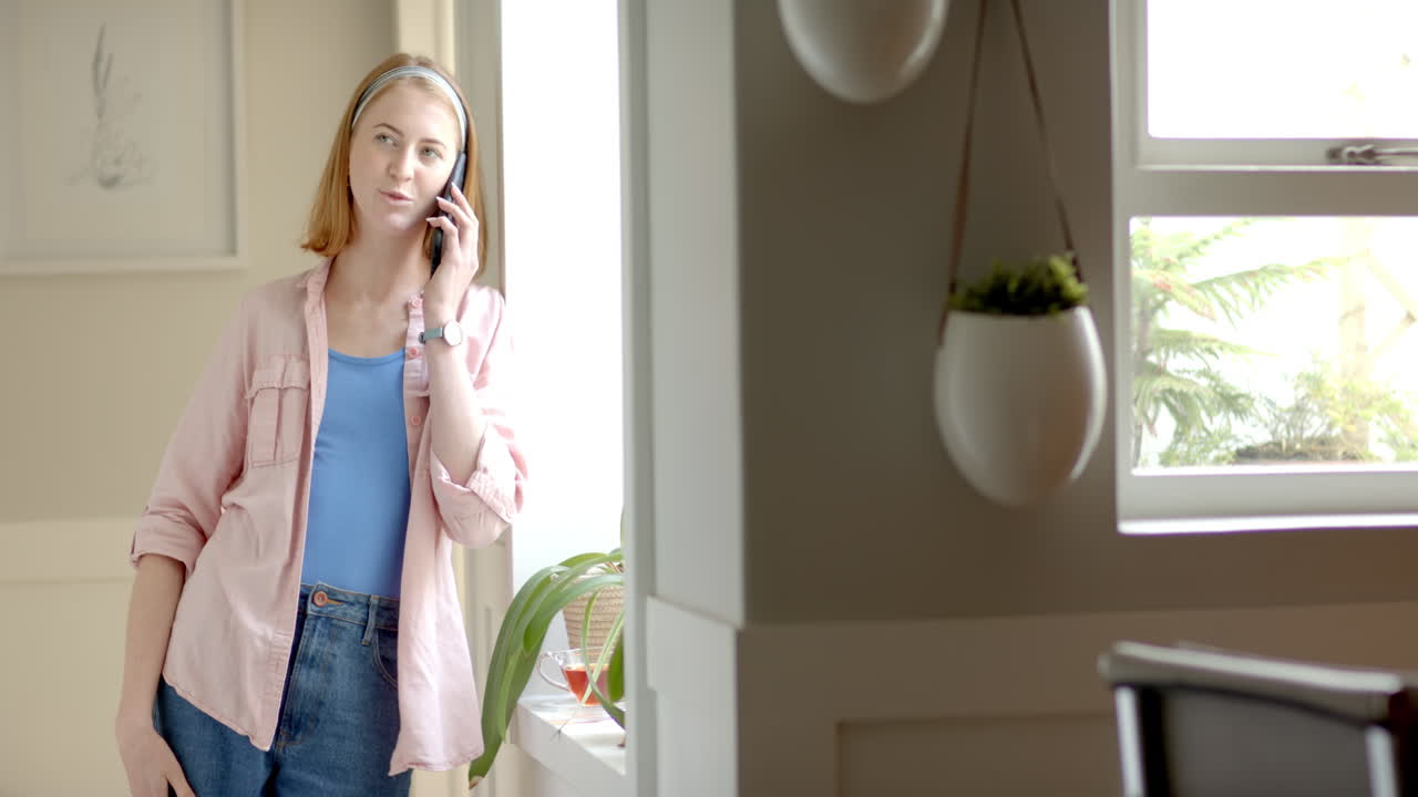Talking on smartphone, woman standing by window in bright home setting