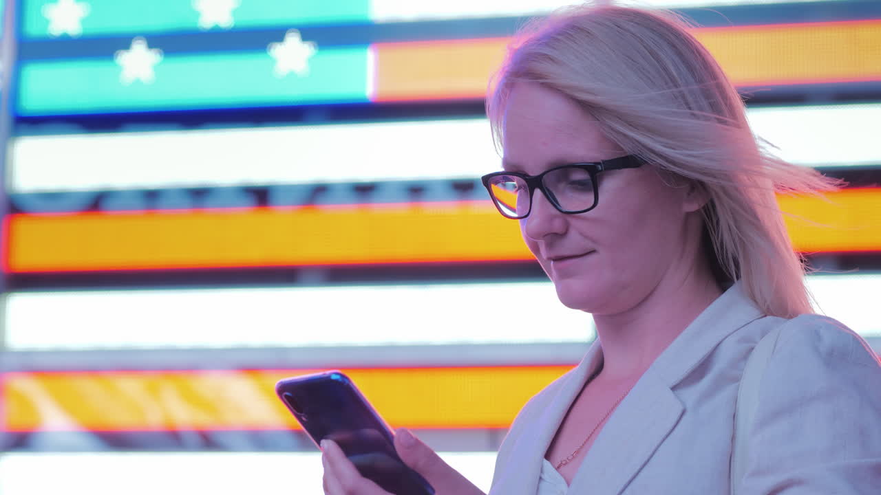 Business Woman Uses A Smartphone On The Background Of A Large American Flag On The Famous Times Squa