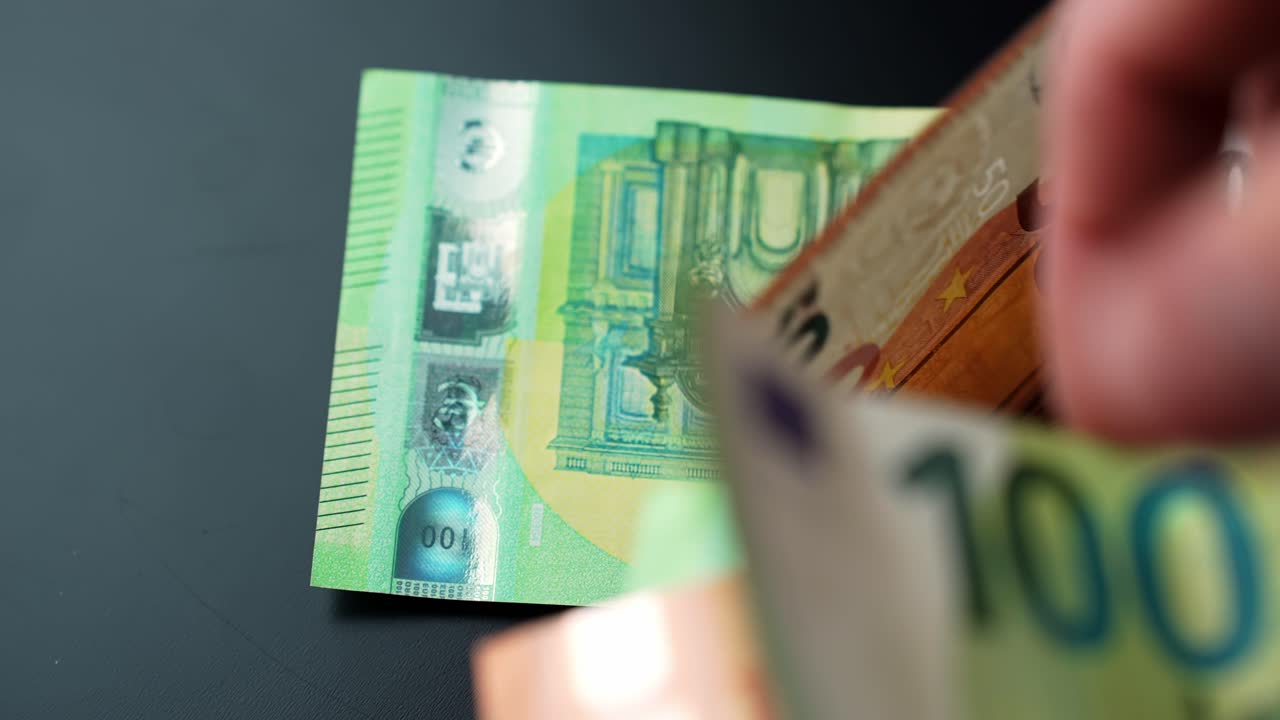 Nærbillede af en hånd, der lægger en 100 euro-pengeseddel oven på en lille bunke på et mørkt bord. Koncept for finans, betaling og budgettering med plads til tekst.