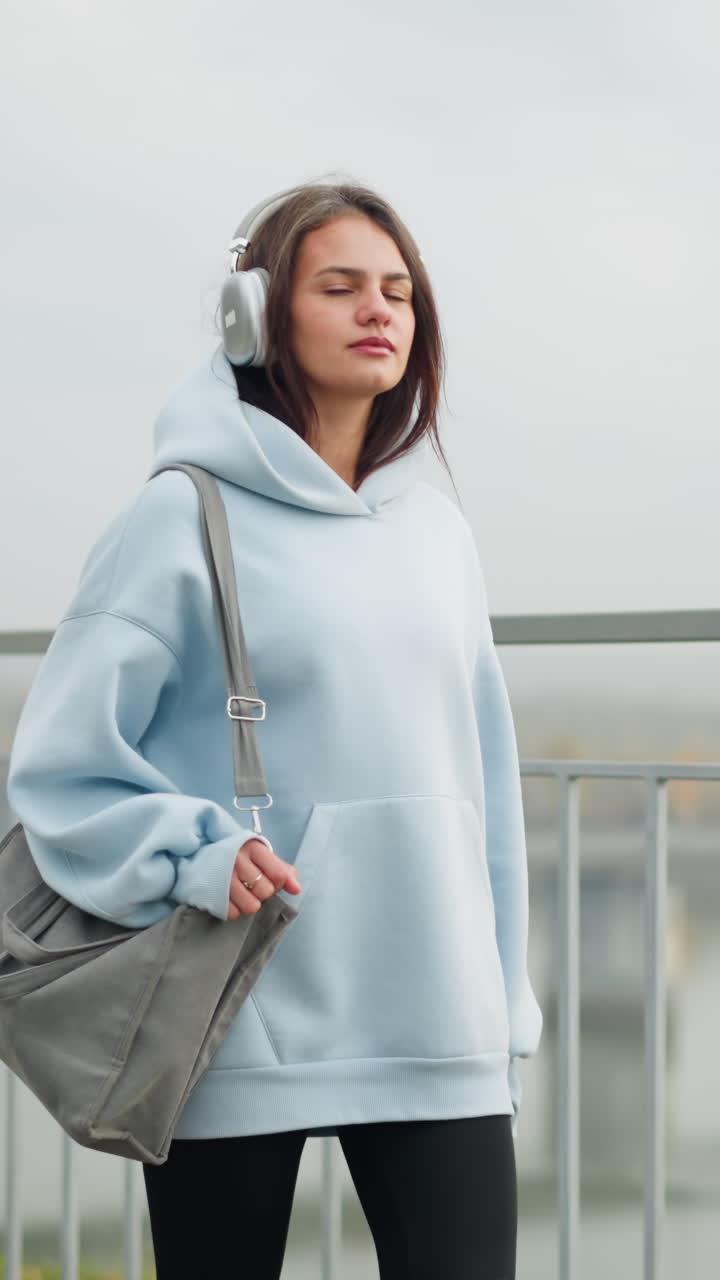 Girl walking confidently with headphones on, holding a bag and a ring in her hand, background features overhead bridge, iron rail, and a river, creating a majestic, urban atmosphere