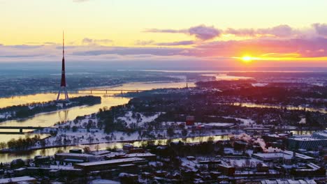 Drone glides left over Riga snowy landscape, revealing colorful winter sunset