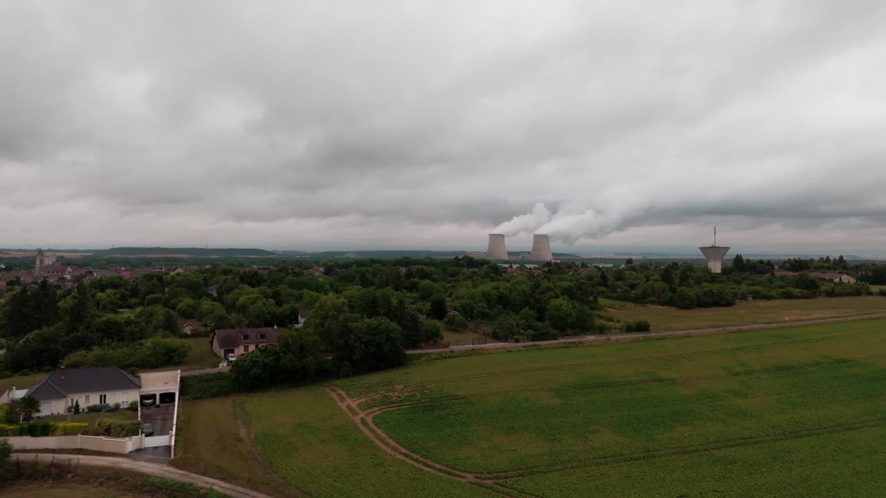 Nuclear power plant, towers emitting steam, rural village and green fields, cloudy sky, France. Aerial forward