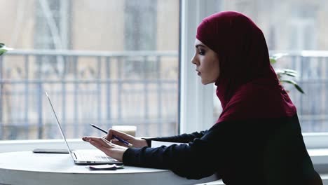 Indoors-footage-of-beautiful-muslim-girl-with-hijab-on-her-head-typing-something-and-then-writing-something-down-in-her-notebook