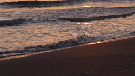 Sandy-beach-at-sunrise-with-waves-breaking-on-shoreline,-reflecting-sunlight,-mediterranean-sea,-close-up,-spain