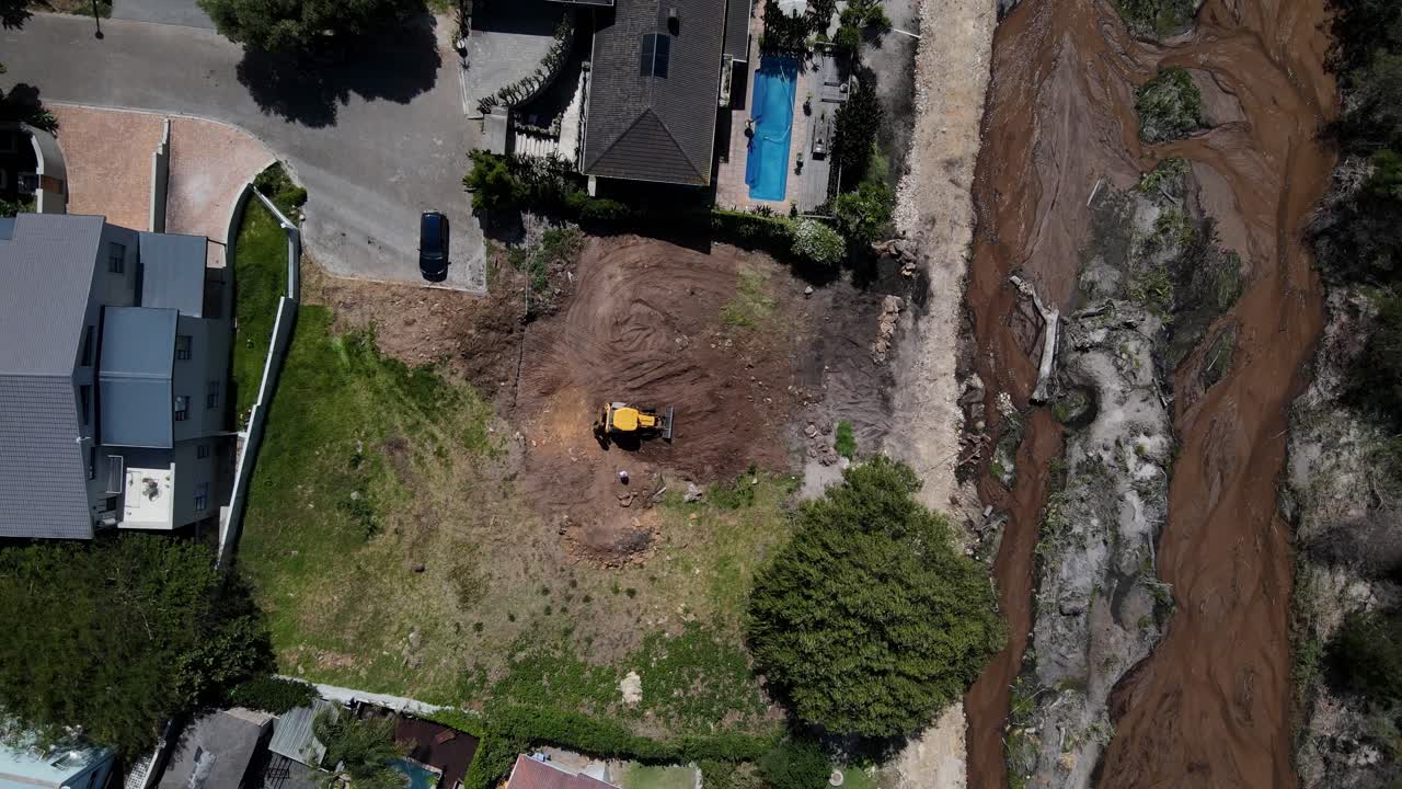 tomada de un avión no tripulado de una excavadora amarilla que despeja un sitio en preparación para la construcción al lado de un río, otras casas y un árbol grande