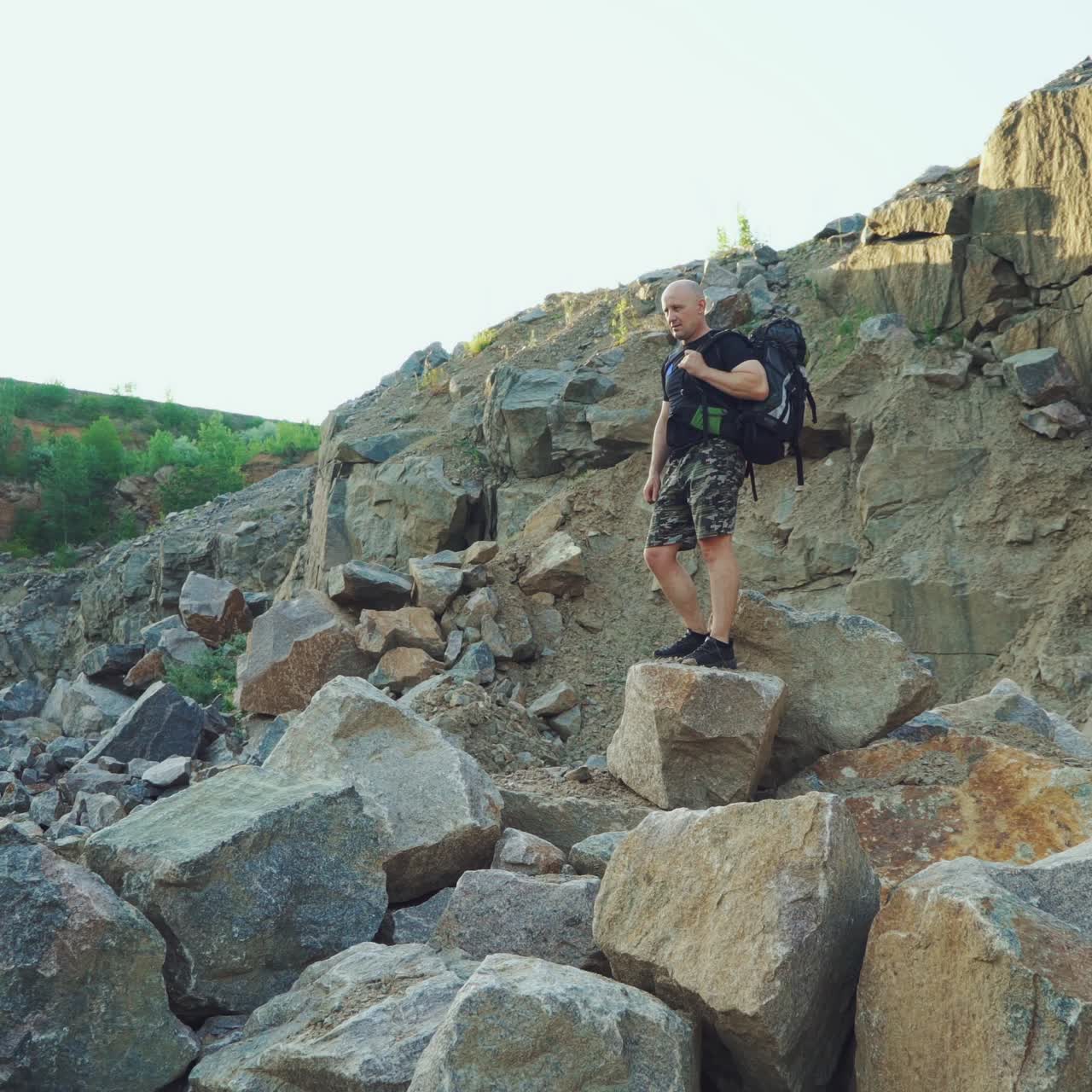 a traveler with a backpack in shorts and a black t-shirt is sitting on a large rock and examining a beautiful landscape around himself