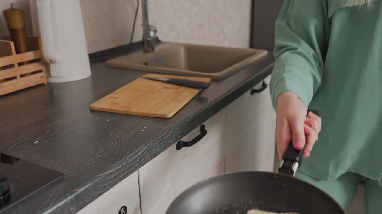 Close up of woman in green pajamas tossing bread slice in frying pan while cooking in modern kitchen, sink, chopping board, knife, and electric kettle visible in background