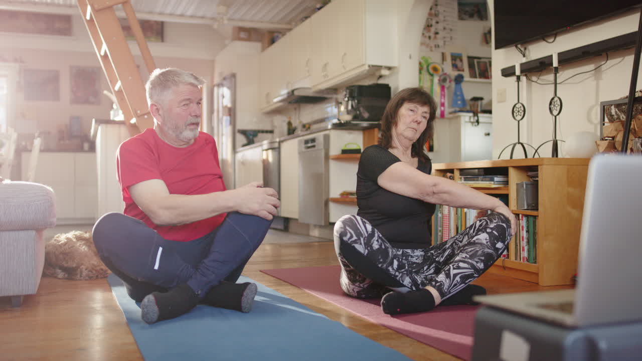 Retired senior married couple sit and follow guided mobility exercises on laptop