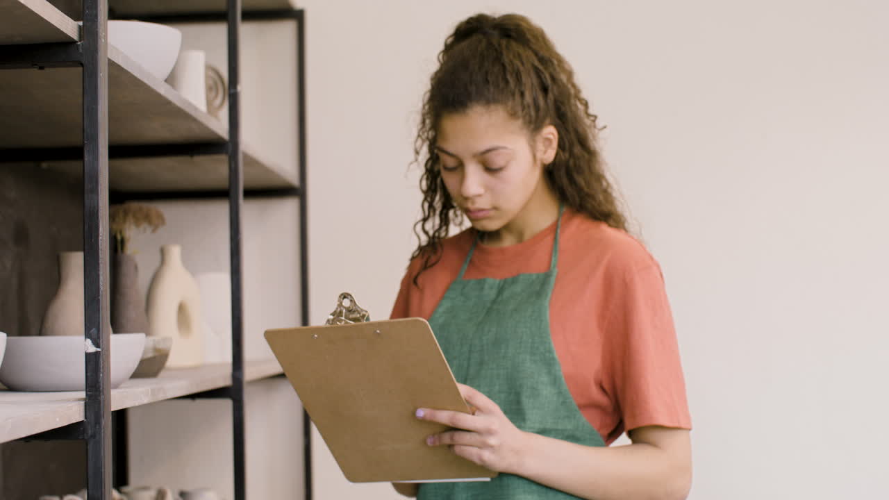 Young Woman Writing On A Clipboard And Making An Inventory Of Ceramics In The Pottery Shop 1