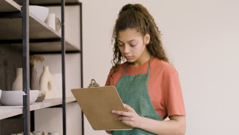 young woman writing on a clipboard and making an inventory of ceramics in the pottery shop 1