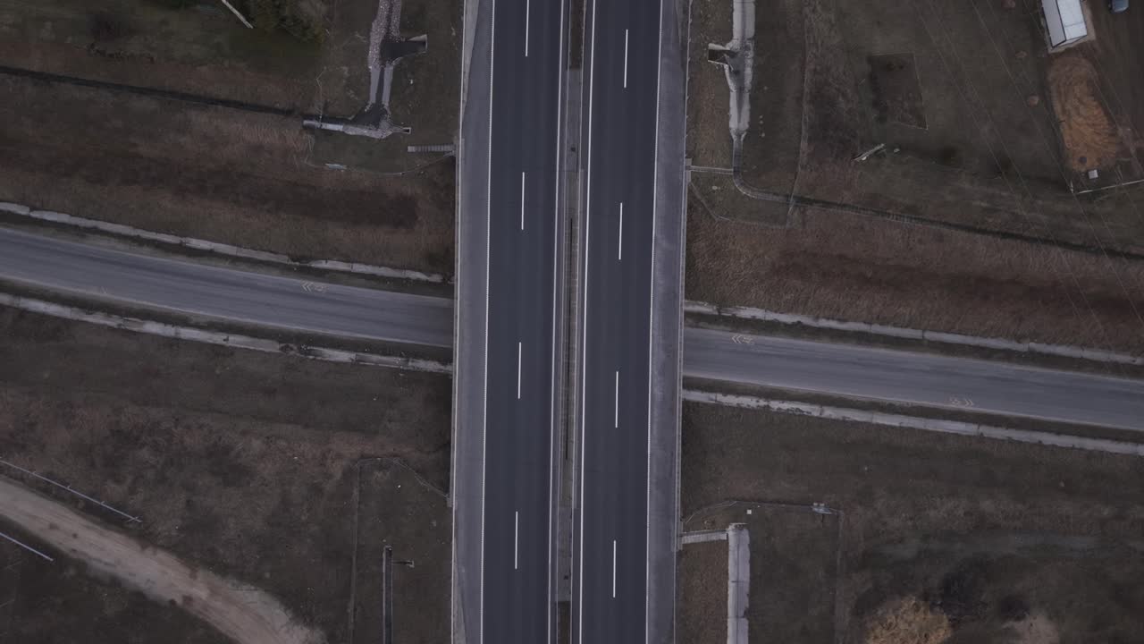 Top down drone view of a highway overpass crossing a smaller rural road. Few cars travel in opposite directions on the multi lane motorway surrounded by dry fields and sparse infrastructure