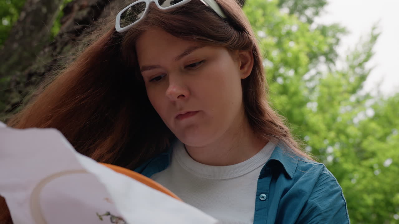 Close up of lady embroidering fabric outdoors with head slanted and hair flowing down, focusing deeply on creative needlework under soft daylight surrounded by green trees