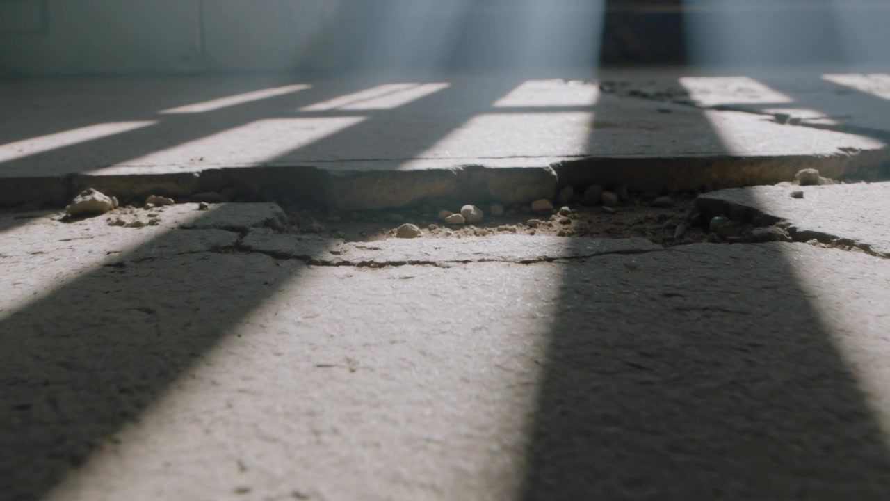 Sliding low camera revealing cracked concrete slab edge at interior, with pebbles, slatted sunlight